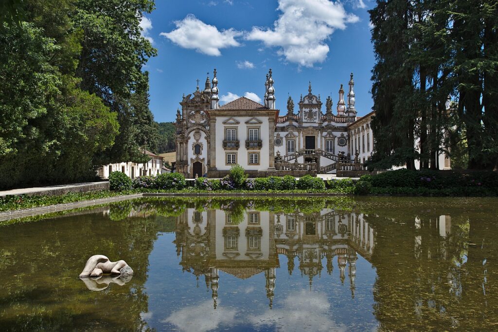 Baroque-style ornate palace reflected in a calm garden pond with lush greenery around.